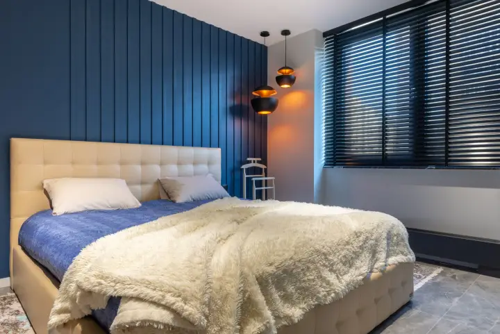 A modern bedroom with a navy panelled wall, pendant lamps and dark horizontal blinds across a wide window.