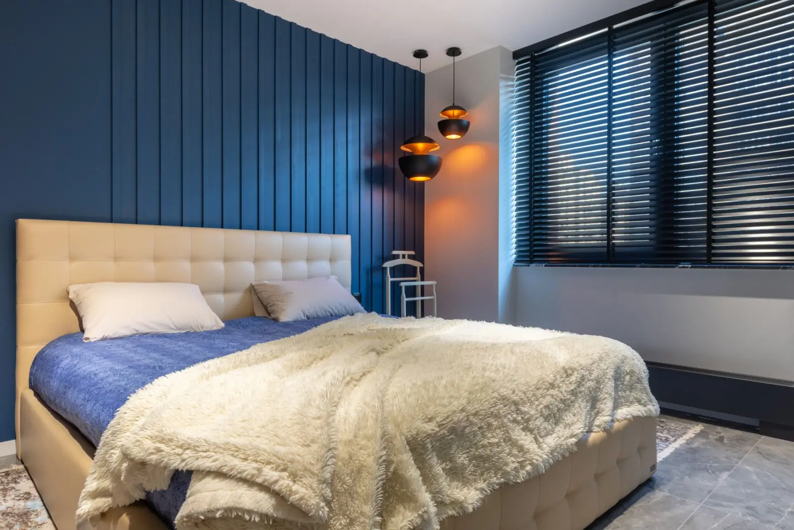 A modern bedroom with a navy panelled wall, pendant lamps and dark horizontal blinds across a wide window.