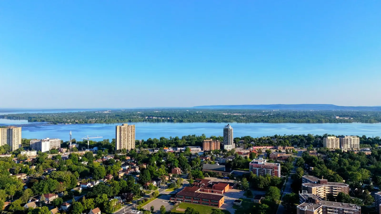 Aerial view over leafy Hamilton neighbourhoods and apartment towers toward the bay and Lake Ontario.