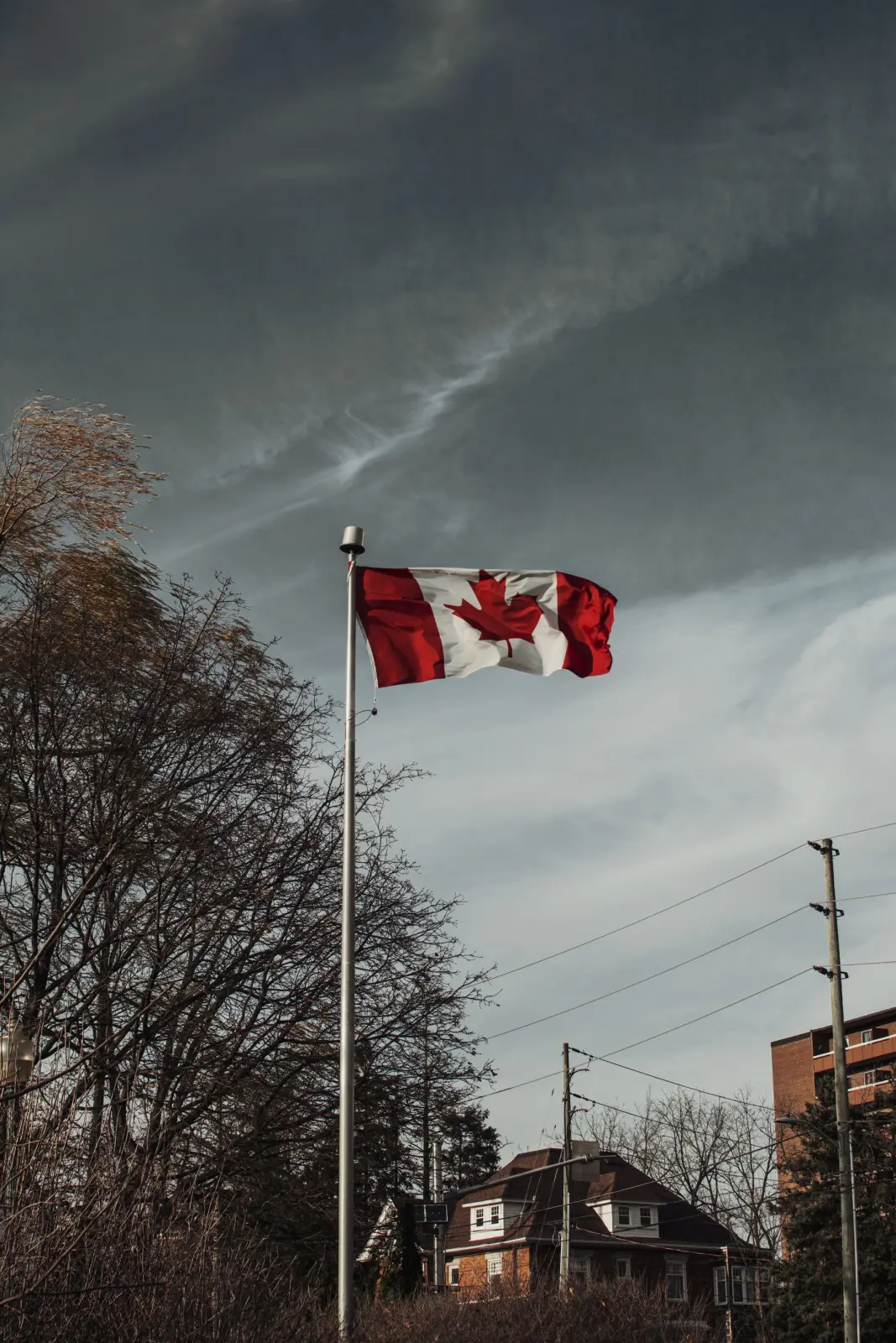 A Canadian flag flies under a moody sky above autumn trees and a brick house in Milton, Ontario.