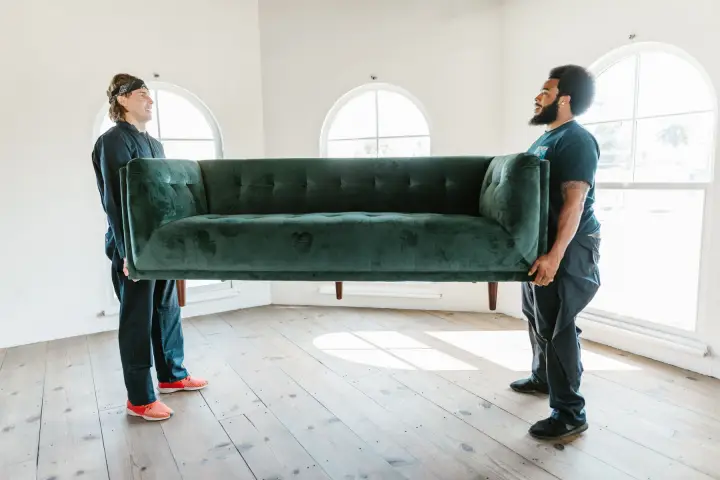 Two movers in dark work clothes carry a green velvet sofa across a bright, empty room with arched windows.