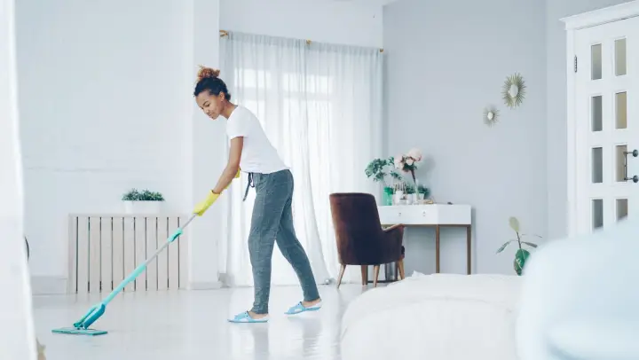 A woman in yellow gloves mops the pale floor of a bright, sparsely furnished room with sheer curtains.