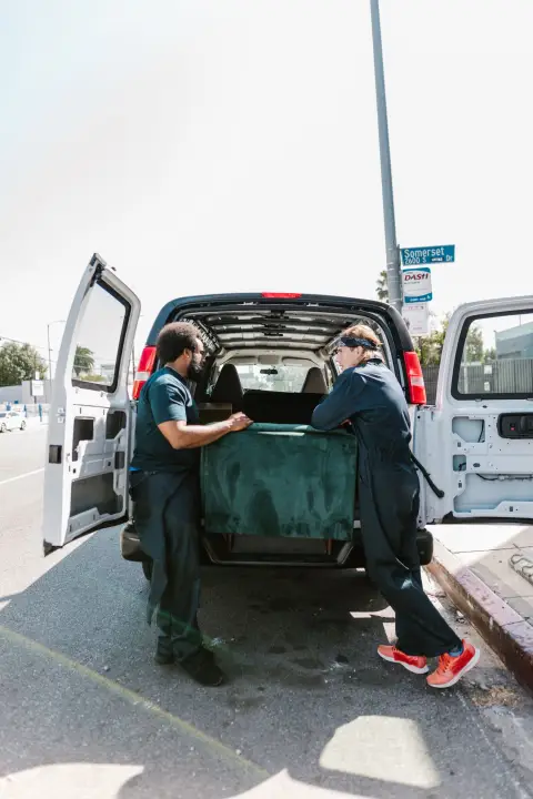 Two movers slide a green sofa into the open back doors of a white van parked at the curb.