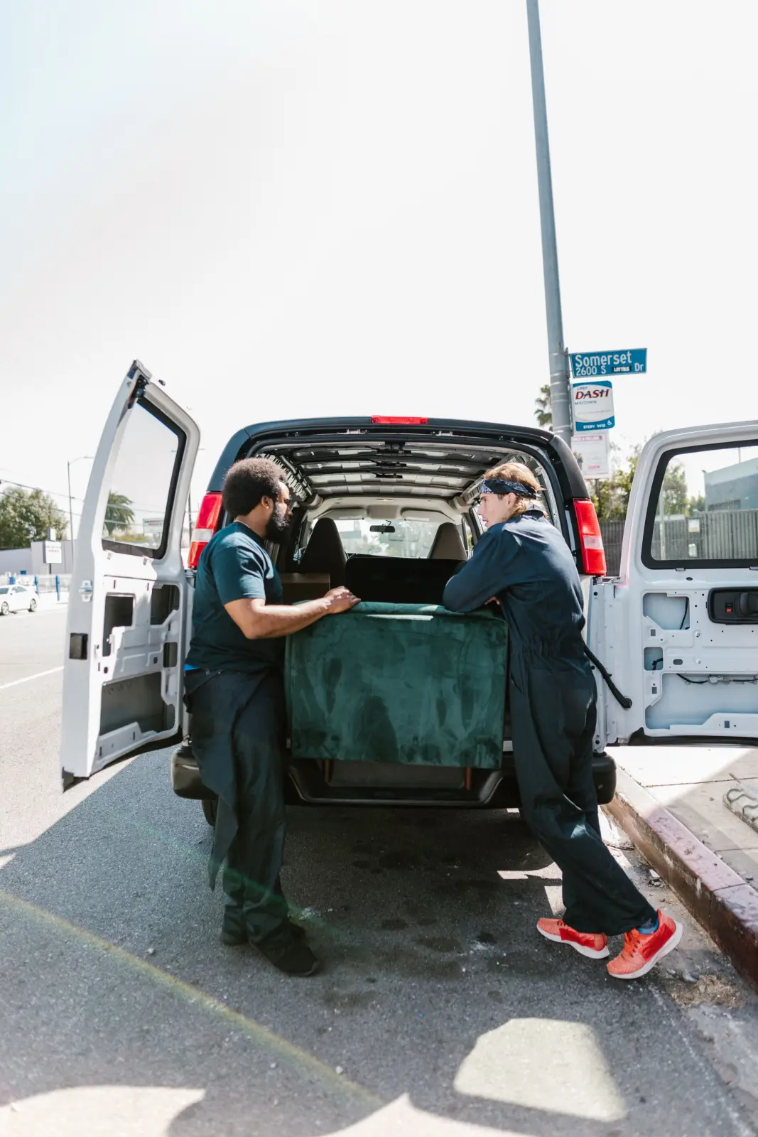 Two movers slide a green sofa into the open back doors of a white van parked at the curb.