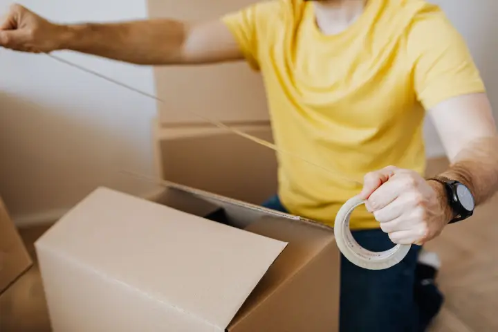 A man in a yellow T-shirt pulls packing tape across a cardboard box, with more boxes stacked behind him.