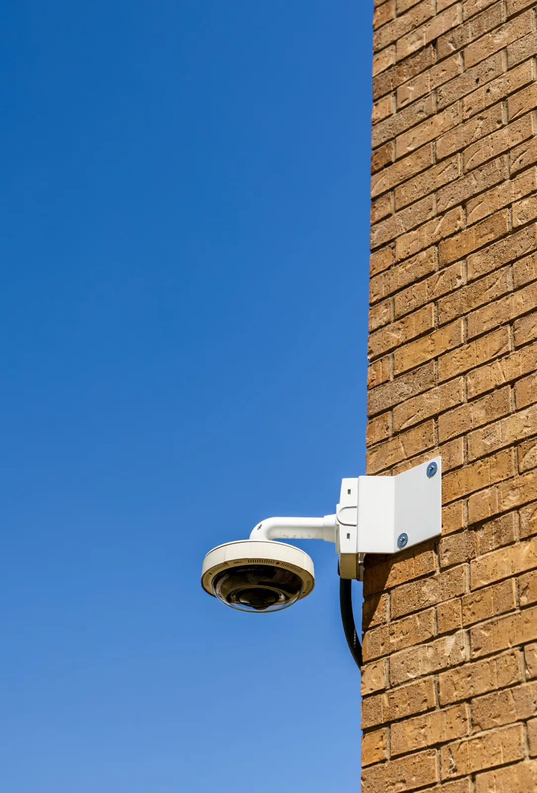 A white dome security camera on a wall bracket fixed to a brick wall, against a clear blue sky.