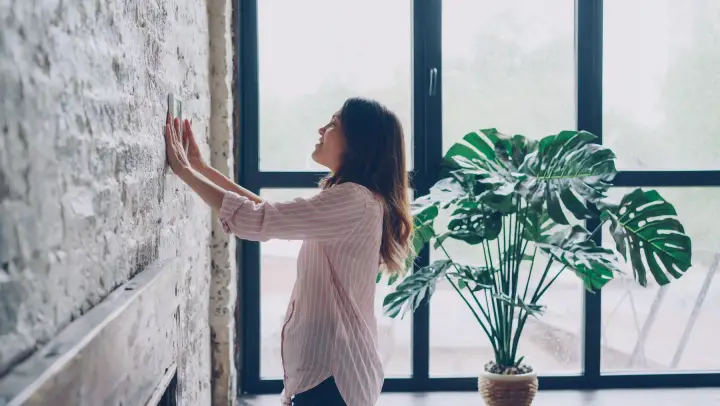 A smiling woman adjusts a wall-mounted thermostat beside a large window and a monstera plant.
