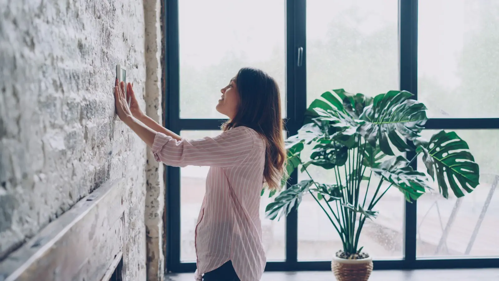 A smiling woman adjusts a wall-mounted thermostat beside a large window and a monstera plant.