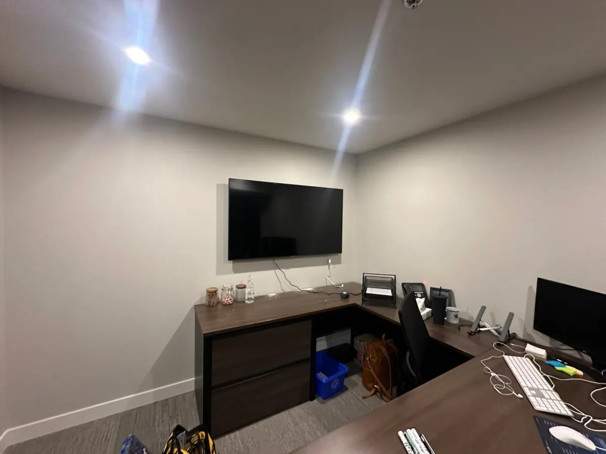 A flat-screen TV wall-mounted above an L-shaped desk in a small office
