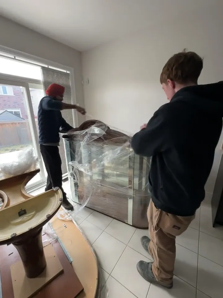 Two crew members wrapping a large wooden furniture piece inside a home